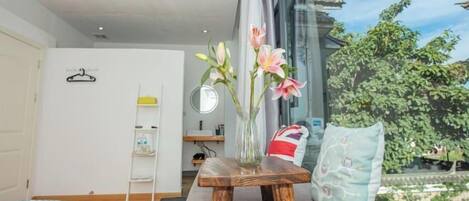 A modern room with a wooden table, a vase with flowers, a window with a view of greenery, and a bed with a blue cover.