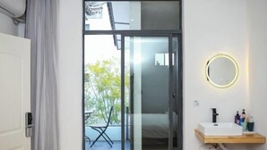 A modern bathroom with a glass door leading to a balcony, a round mirror, and a wooden shelf with toiletries. - Erduobian Inn (Dali City)