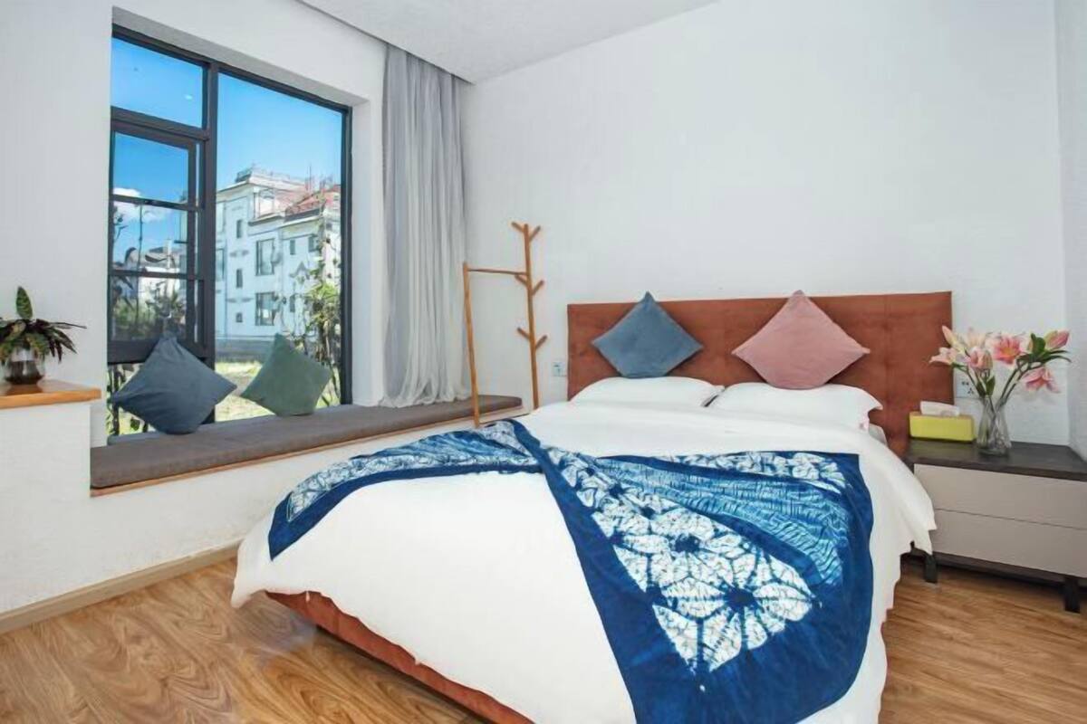 A bedroom with a large window, a bed with a blue and white patterned bedspread, a wooden headboard, and a bedside table with a vase of flowers.