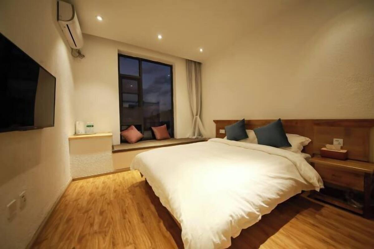 A hotel room with a large bed, a bedside table, a window with curtains, and an air conditioning unit.