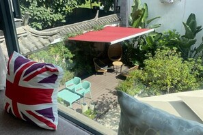 Uitzicht vanuit een raam op een terras met een rode parasol, tuinmeubilair en weelderige beplanting.