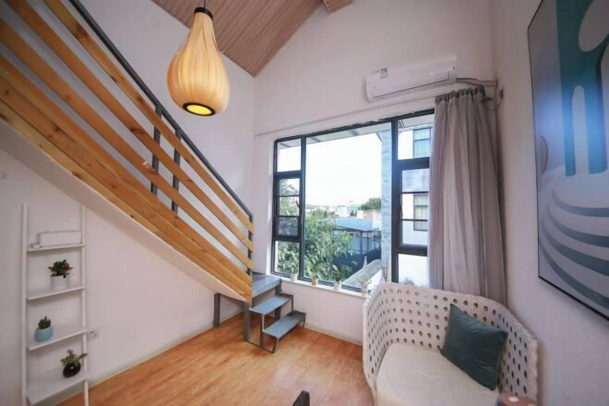 A room with a wooden staircase, a white sofa with a patterned cover, a large window with a view, and a hanging light fixture.