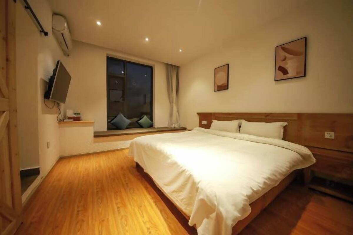 A bedroom with a large bed, a window with curtains, a mounted TV, and a wooden floor.