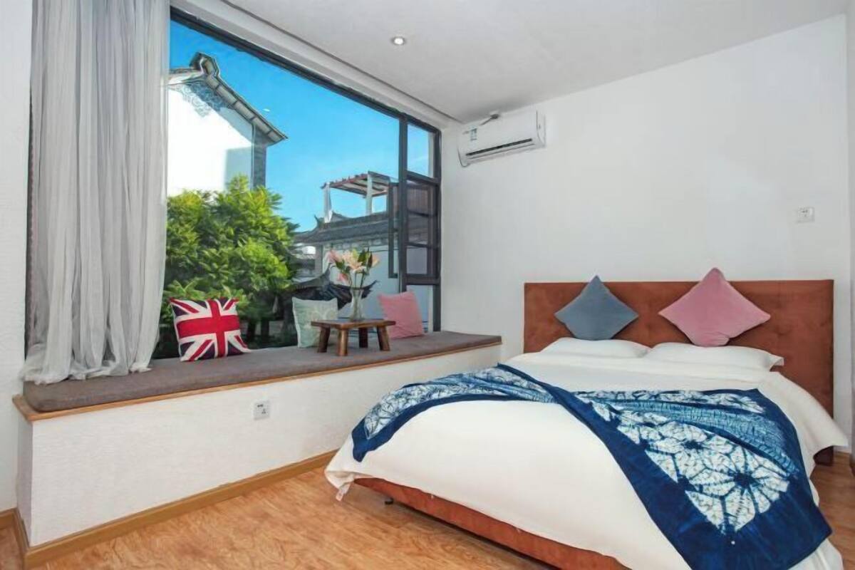 A bedroom with a large window, a bed with a blue and white patterned bedspread, and a view of a building and trees outside.