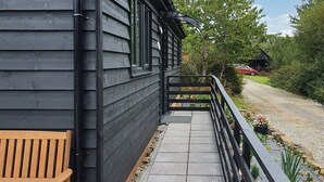 A wooden building with a balcony, a bench, and a gravel path leading to a red car. - Fern Lodge - UK40018 (Halwill Junction, near Beaworthy)