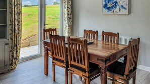 A dining area with a wooden table and chairs, a view of a green field through a glass door, and a framed artwork on the wall. - Fern Lodge - UK40018 (Halwill Junction, near Beaworthy)