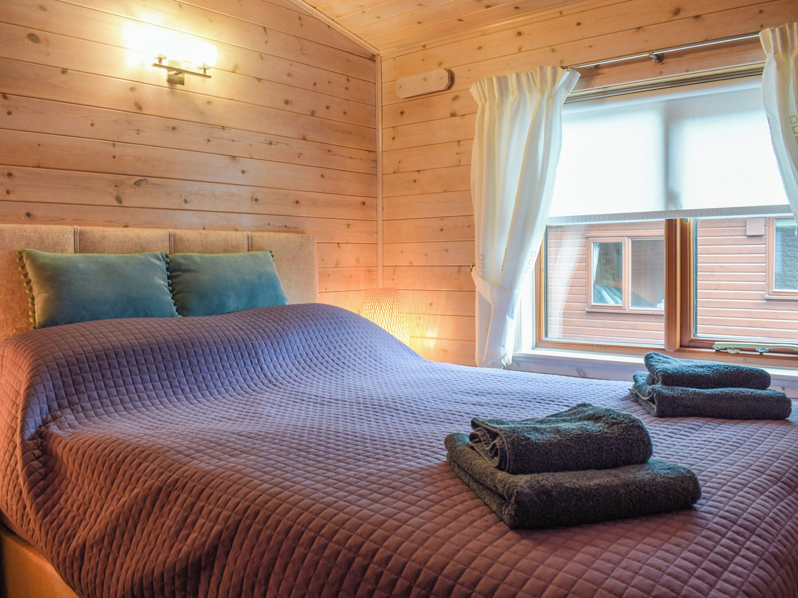 A wooden cabin room with a bed, pillows, and a window with curtains.