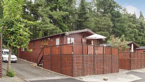 A wooden cabin with a fenced patio area, a car parked outside, and a forested backdrop. - 2 bedroom accommodation in Bridgnorth (Bridgnorth)