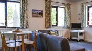 Een gezellige woonkamer met een bank, een eettafel met stoelen, een televisie op een dressoir, en grote ramen met gordijnen.
