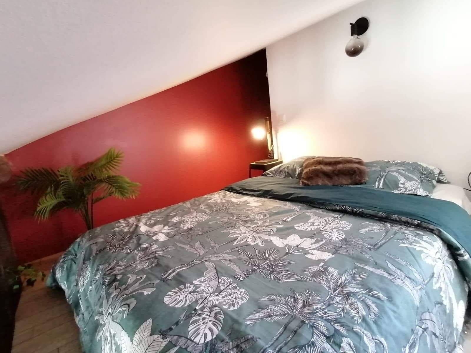 Slaapkamer met een rode accentmuur, een bed met een bloemenprintendekbed, een nachtkastje, een lamp en een kleine plant.