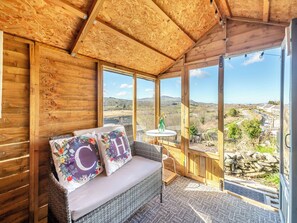 A wooden interior with a wicker sofa, a small table with a vase, and large windows offering a view of the countryside. - 2 bedroom accommodation in Talysarn (Talysarn)