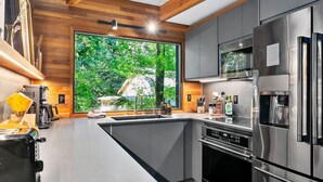 A modern kitchen with stainless steel appliances, a large window with a view of greenery, and wooden paneling on the walls.