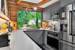 A modern kitchen with stainless steel appliances, a large window with a view of greenery, and wooden paneling on the walls.