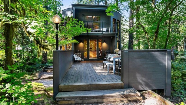 A modern two-story cabin with a deck, surrounded by dense forest.