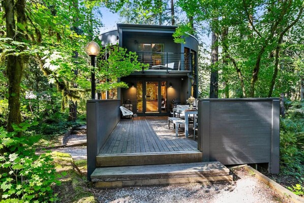A modern two-story cabin with a deck, surrounded by dense forest.