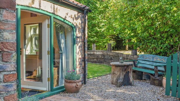 Een stenen huis met een groene deur, een houten bank en een tafeltje met een beeldje.