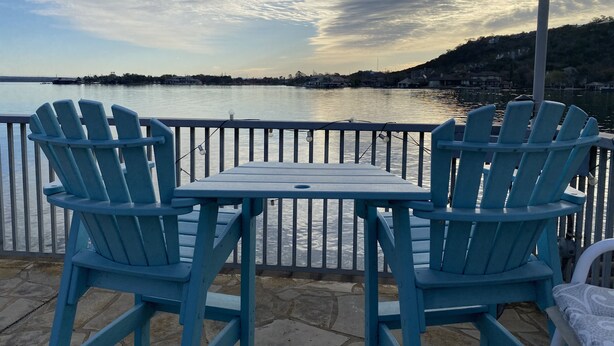Zwei blaue Adirondack-Stühle und ein kleiner Tisch auf einer Steinterrasse mit Blick auf ein Gewässer und eine entfernte Uferlinie.