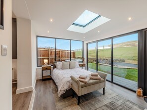 A bedroom with a skylight, large windows, a bed, bedside tables, and a view of a green landscape.