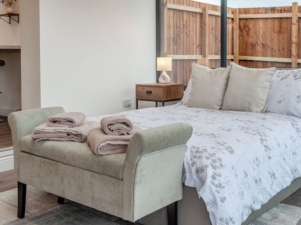 Een slaapkamer met een bed, nachtkastje, lamp, stoel en uitzicht op een houten schutting.