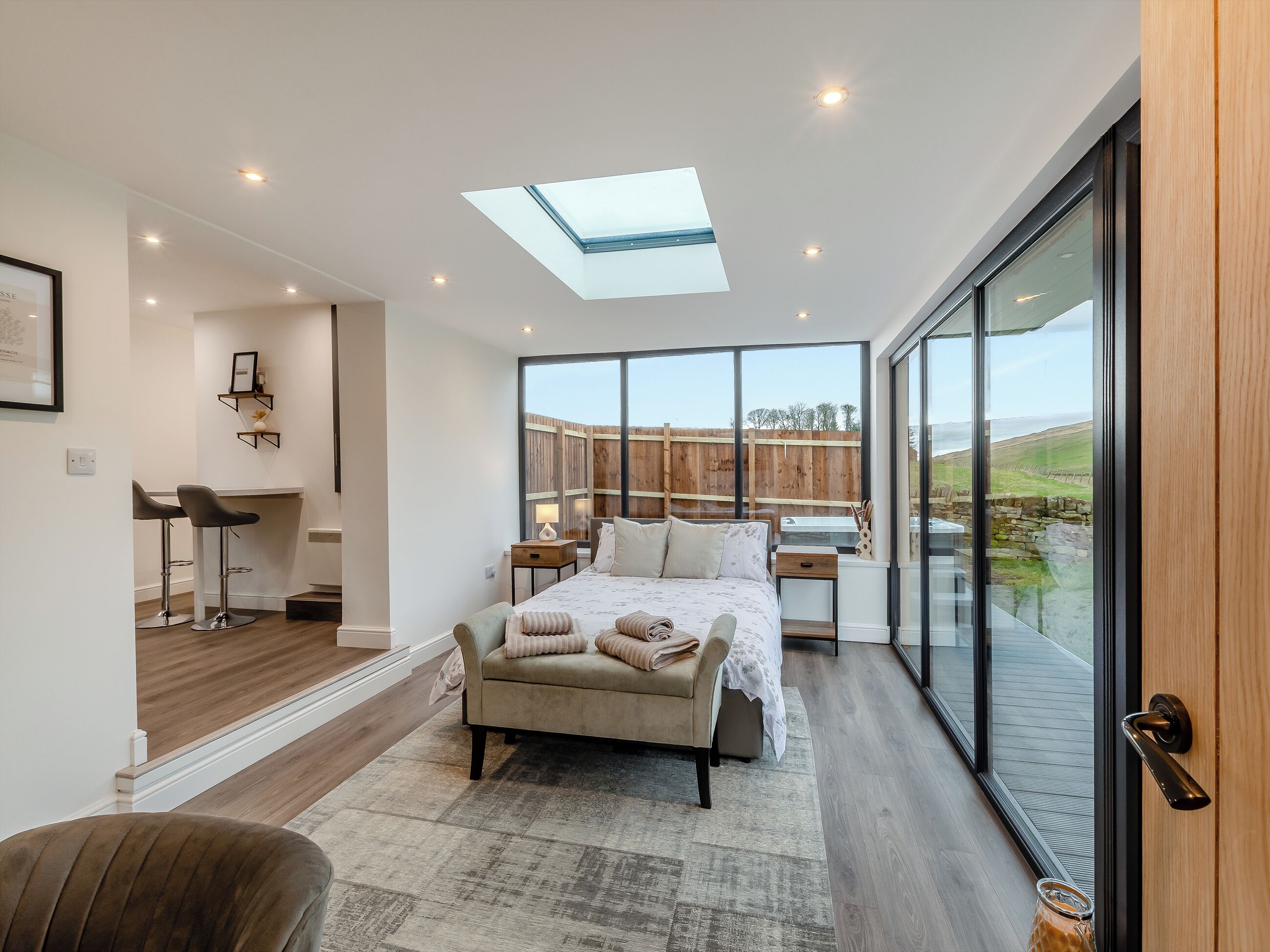 A bedroom with a skylight, large windows, a bed, bedside tables, and a view of the outdoors.