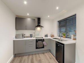 Een moderne keuken met grijze kastjes, een inbouwoven, een vaatwasser en een raam met zonwering.