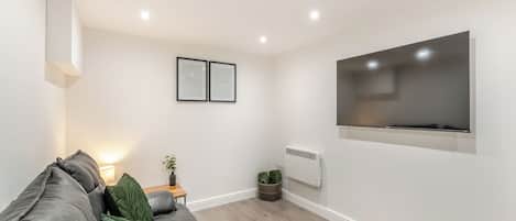 Un salón moderno con un sofá gris, un televisor de pantalla plana, diplomas enmarcados en la pared y una mesita con una planta.