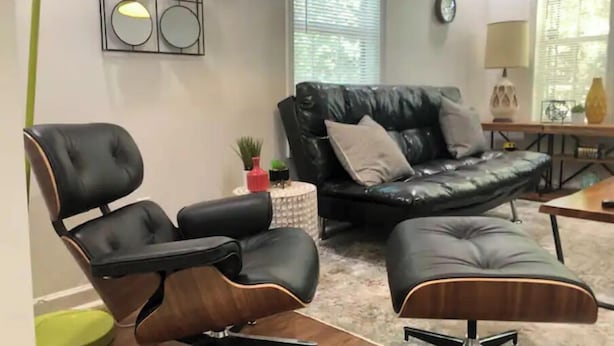 A modern living room with a black leather sofa, a black leather armchair, and a matching ottoman. There is a small round table with a plant and a red vase. A wall-mounted light fixture with circular mirrors is visible. - “Mint City w Soul...” 2BR 5 min to Stadium & UMMC! (Jackson)