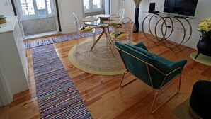 Un salon moderne avec une table à manger en verre, des chaises transparentes, un canapé vert, un téléviseur posé sur un meuble circulaire et une grande fenêtre donnant sur des immeubles.