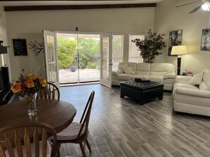 A spacious living room with a wooden dining table, two white sofas, a black ottoman, and a large window with a view of a garden.