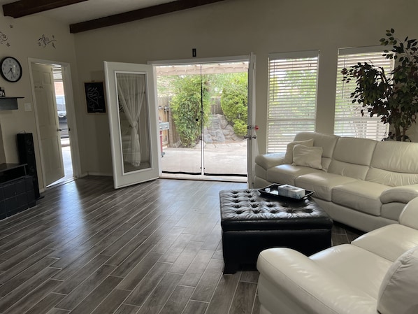 A living room with a white leather sofa, a black leather ottoman, and a wooden floor. There is a sliding glass door leading to a patio with a view of greenery.