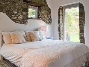 A bedroom with a stone wall, a large bed with multiple pillows, a nightstand with a lamp, and a view through a window.