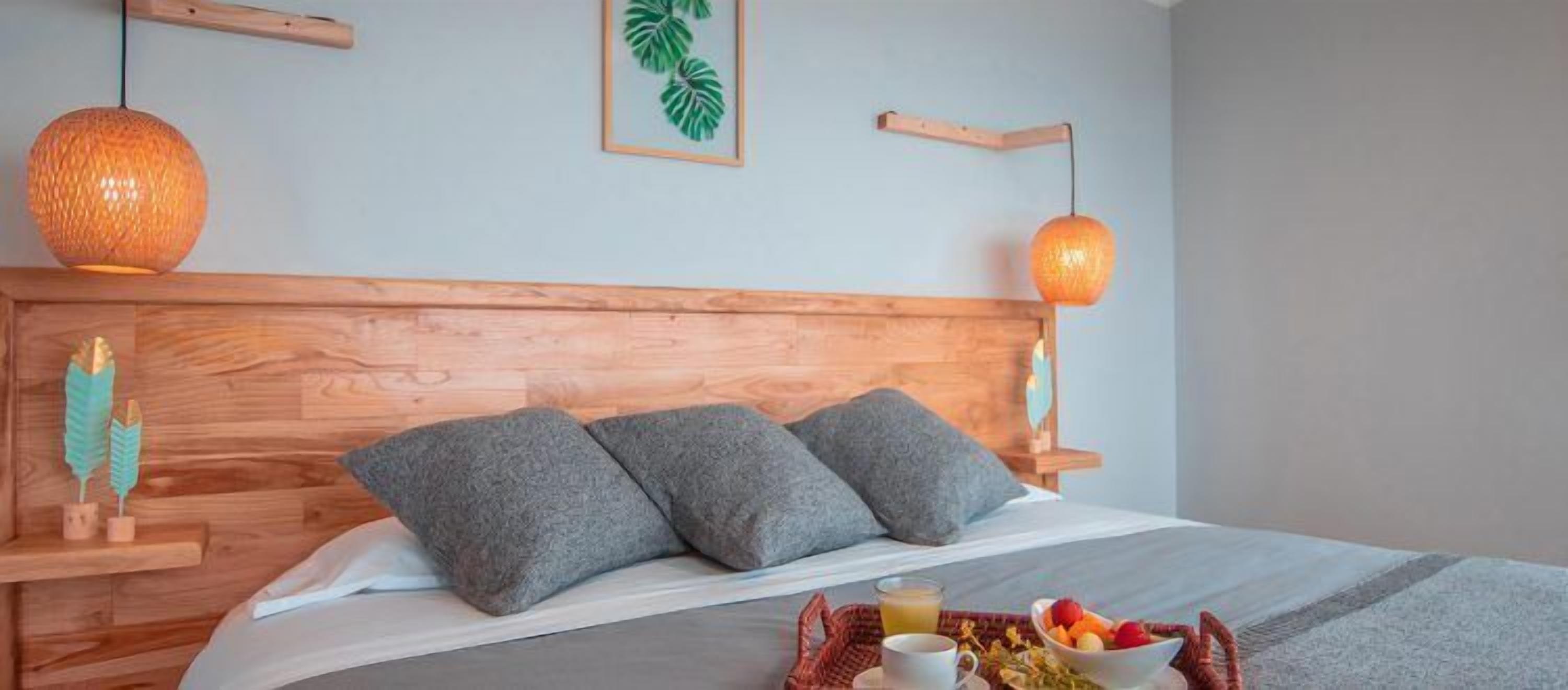 A neatly made bed with grey pillows, a wooden headboard, and a fruit basket on the bedside table.