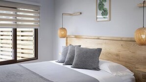 Un dormitorio con cabecera de madera, cama con ropa de cama gris, dos lámparas de noche y una ventana con persianas.