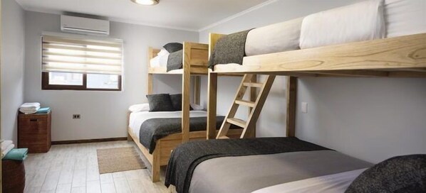 A dormitory room with bunk beds, a window with blinds, a small wooden cabinet, and a floor lamp.