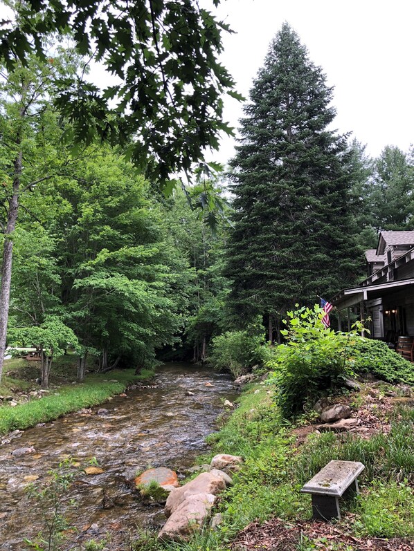 A serene riverside setting with a wooden cabin, lush greenery, and a small bench. - Cabin 4 - Off the grid cabin at Pioneer Village (Maggie Valley)
