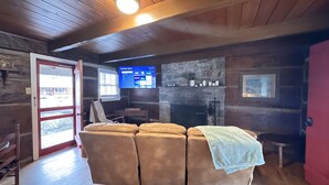 A cozy cabin interior with a wooden ceiling, a stone fireplace, a leather sofa, and a television displaying a menu. - Cabin 4 - Off the grid cabin at Pioneer Village (Maggie Valley)
