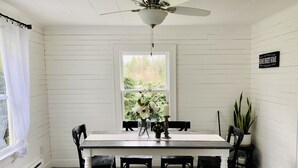 Een eetruimte met een houten tafel, zwarte stoelen, een bank, een plafondventilator, een raam met uitzicht op groen, en een bord aan de muur.