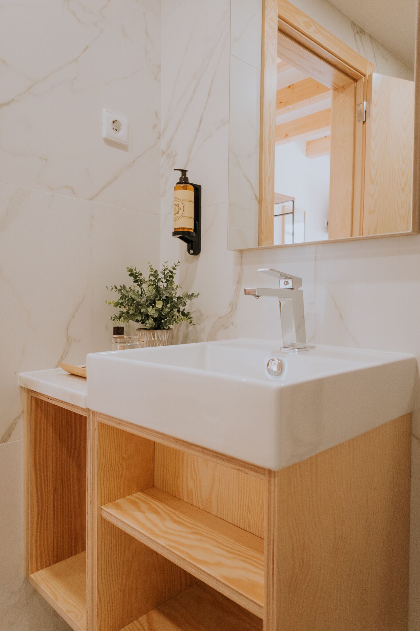Un baño con pared de mármol blanco, tocador de madera, grifo moderno, una planta pequeña e iluminación empotrada.