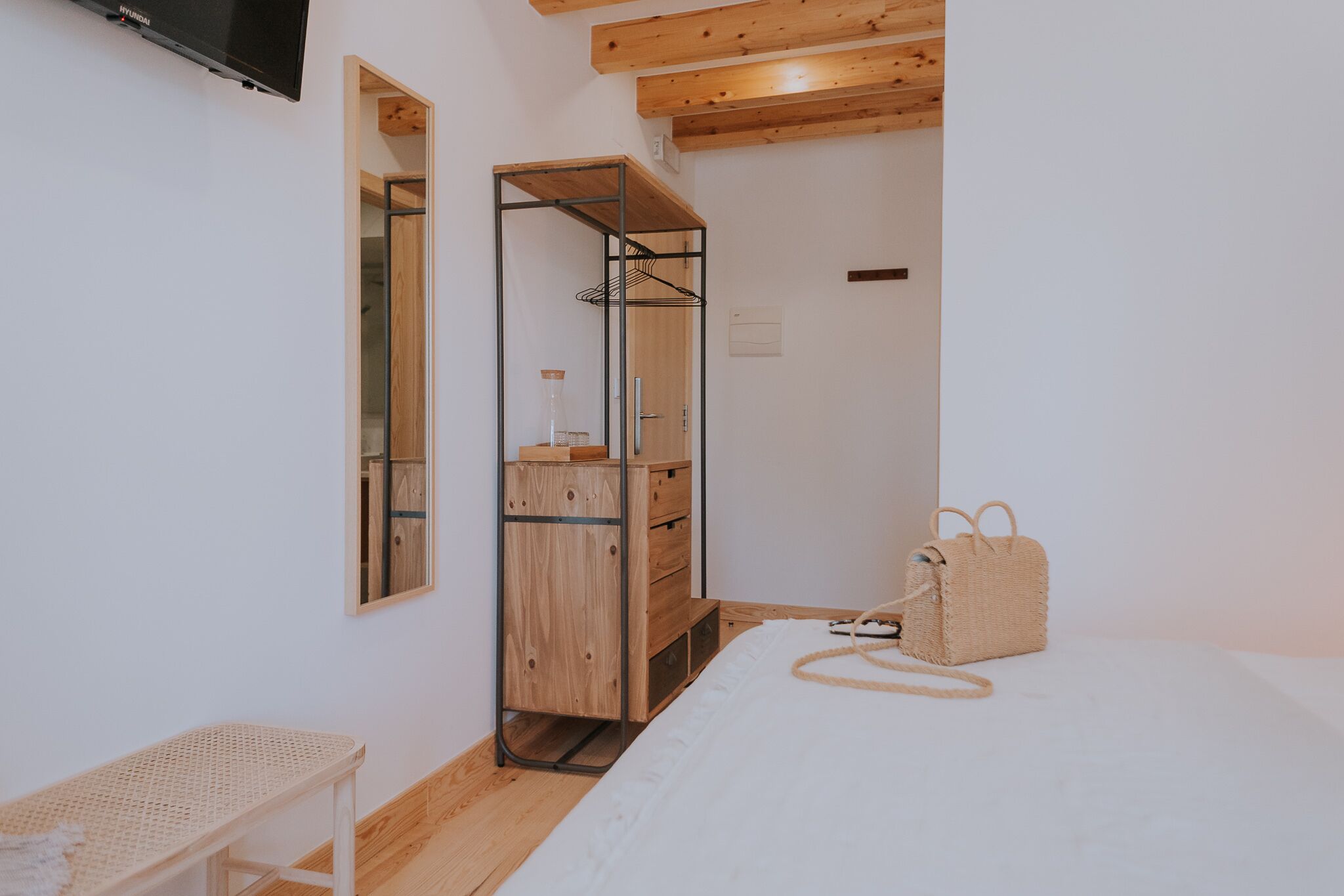 A bedroom with a bed, a wooden dresser, a mirror, a hanging rack, and a wicker handbag.