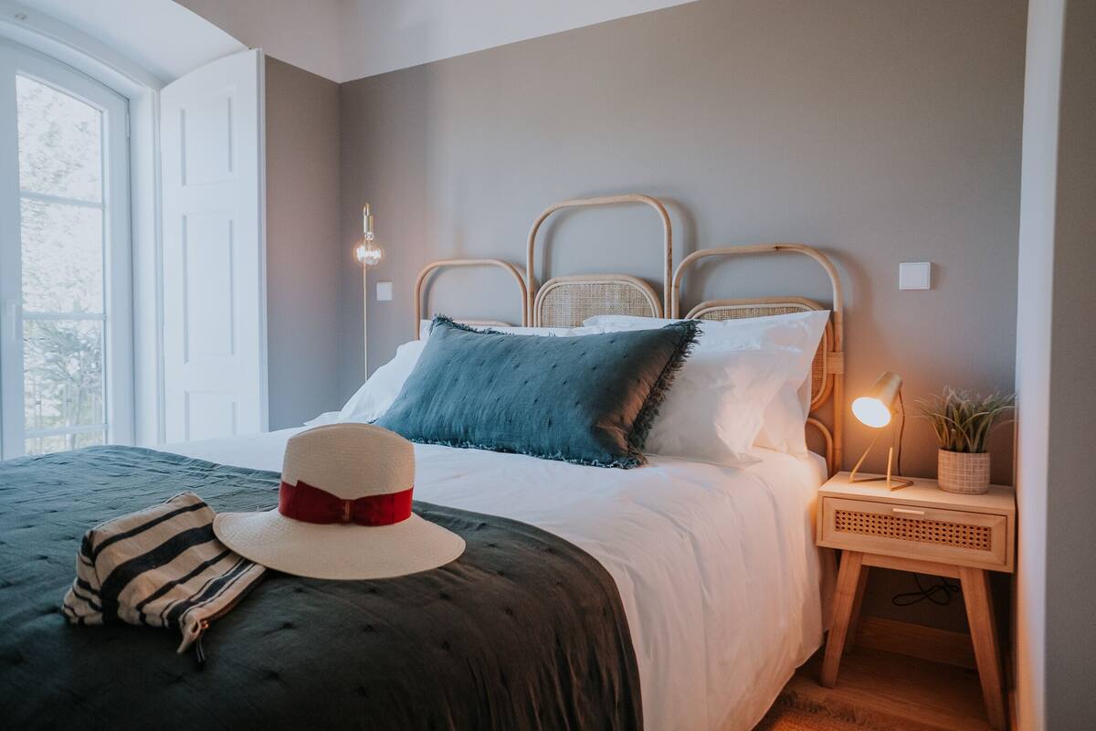 A neatly made bed with a striped pillow, a hat, and a bedside lamp.