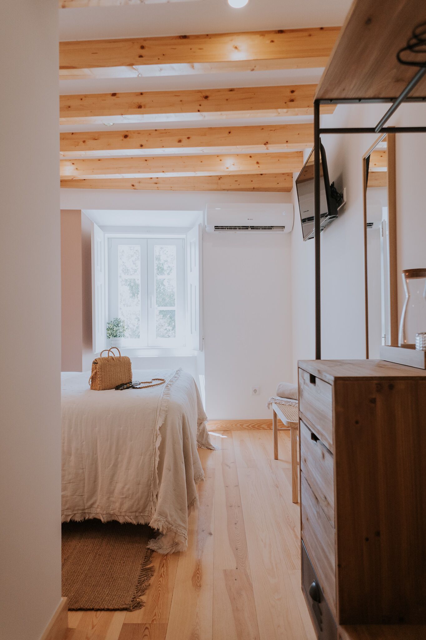 A bedroom with a wooden ceiling, a bed with a white bedspread, a wooden dresser, and a window with a view of trees.