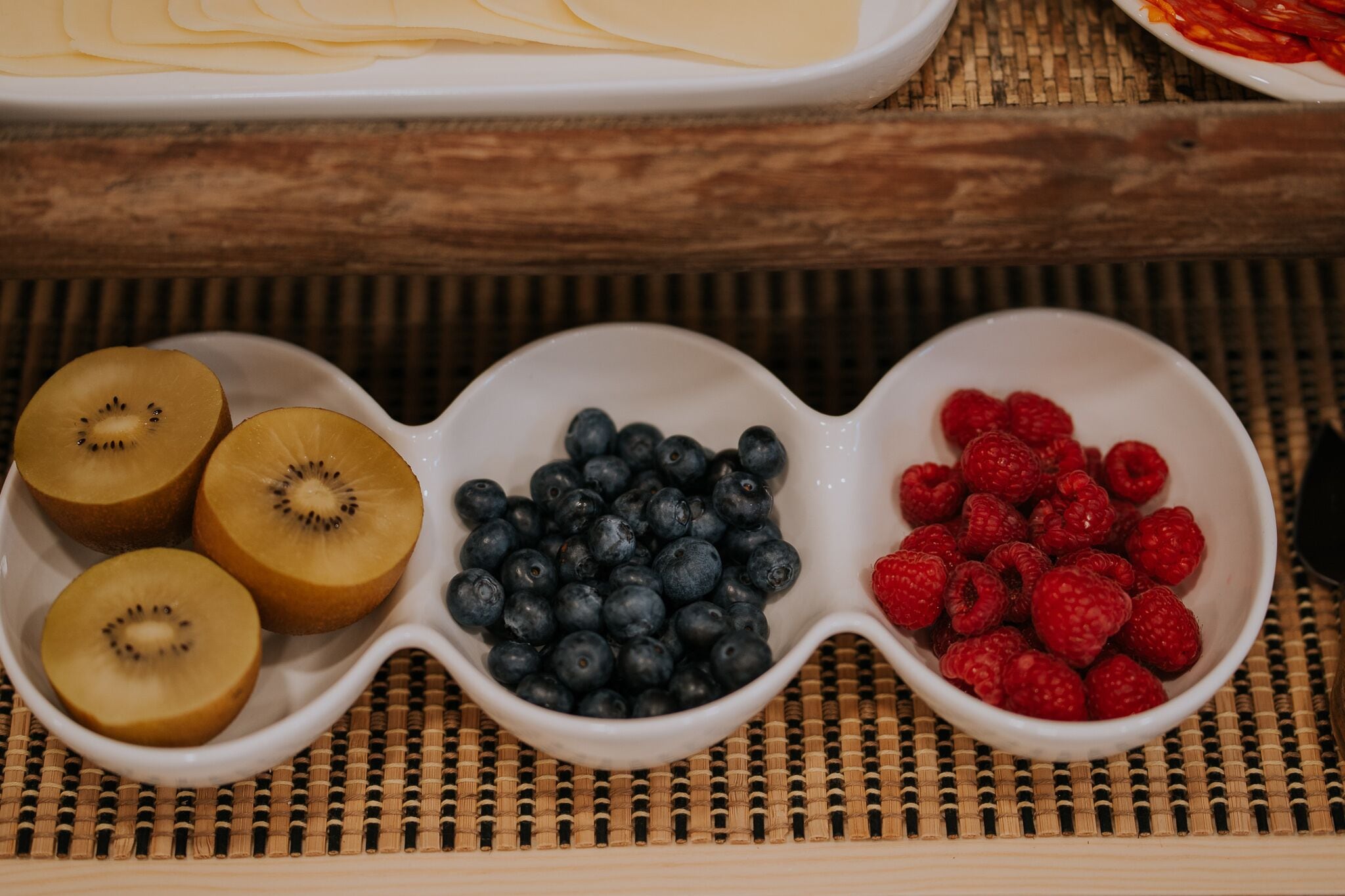 Three white bowls containing sliced kiwi, blueberries, and raspberries on a woven mat.