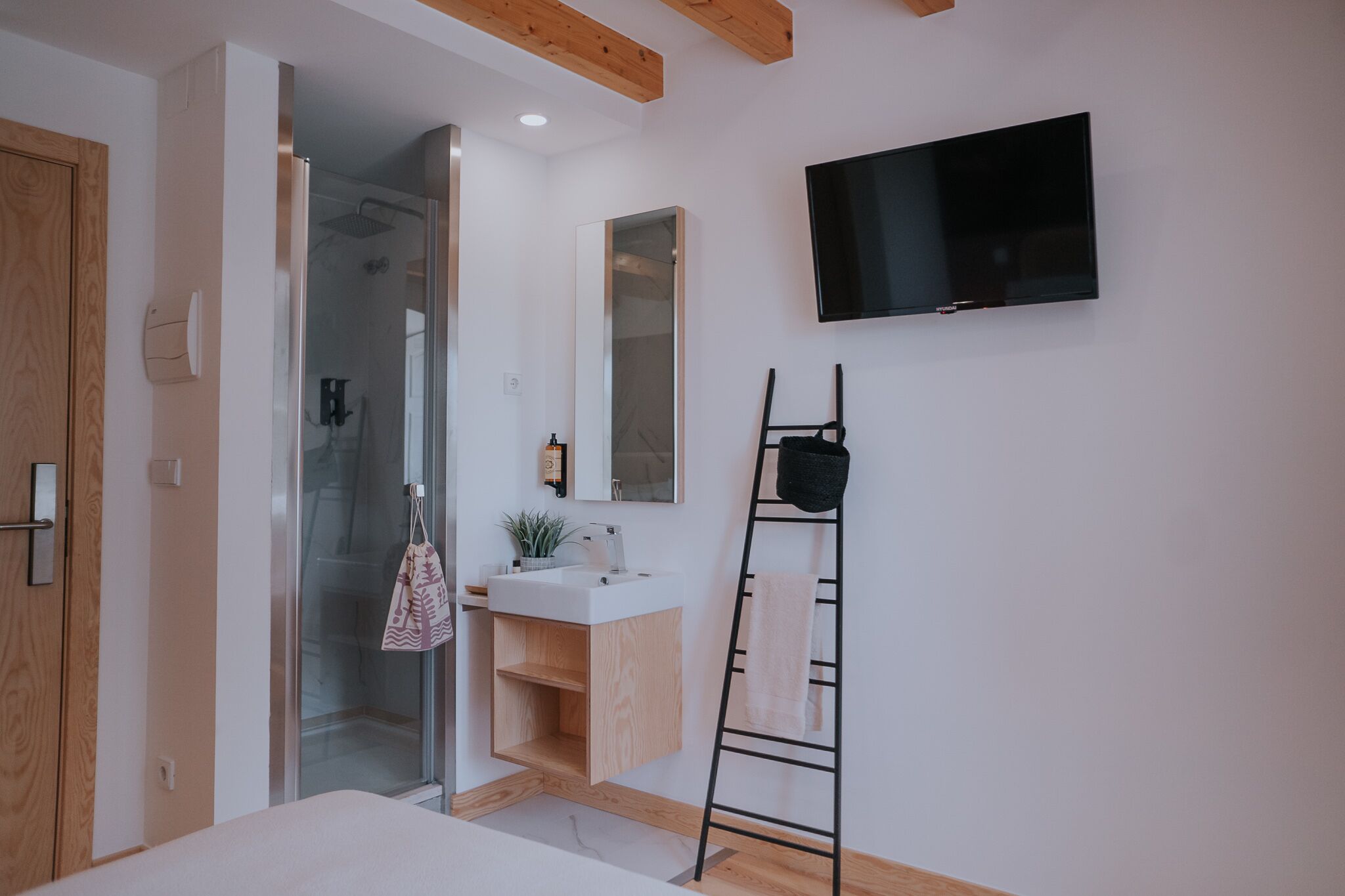 A modern bathroom with a glass shower, a white sink, a wooden vanity, a towel rack, and a mounted TV.