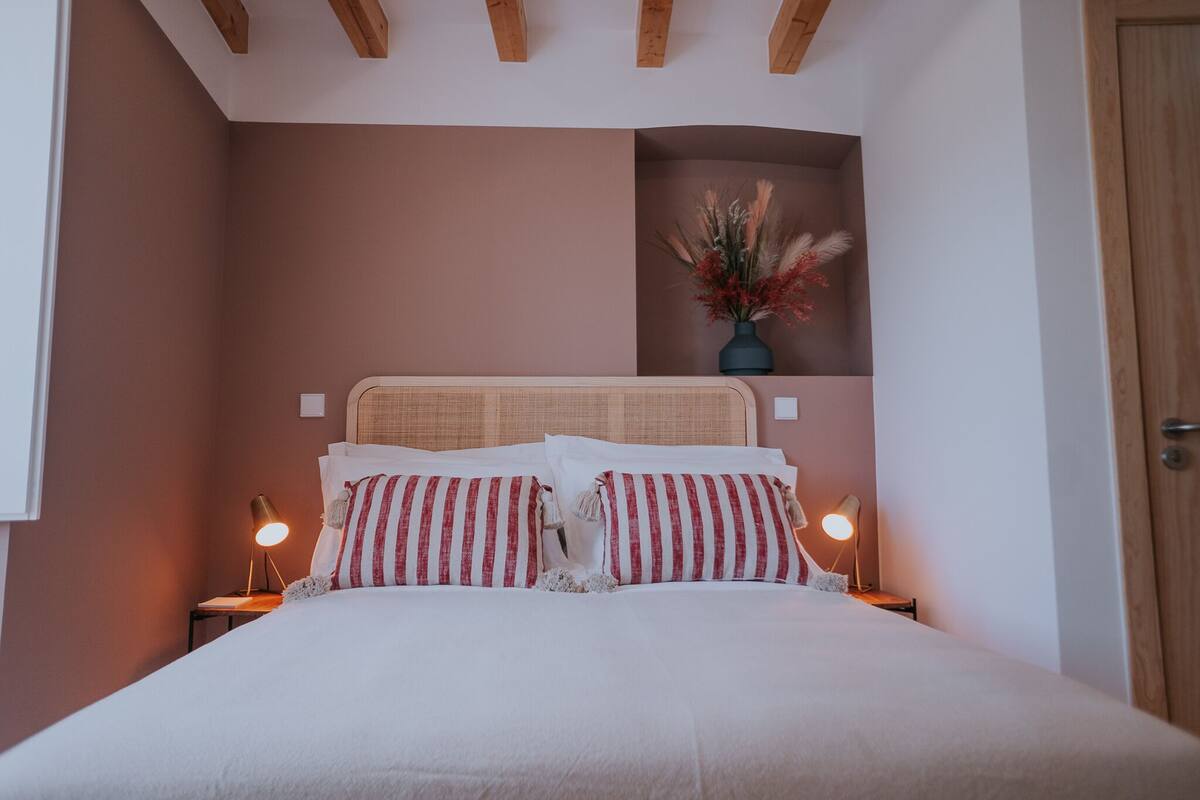 A neatly made bed with striped pillows, two bedside lamps, and a vase with dried flowers on a bedside table.