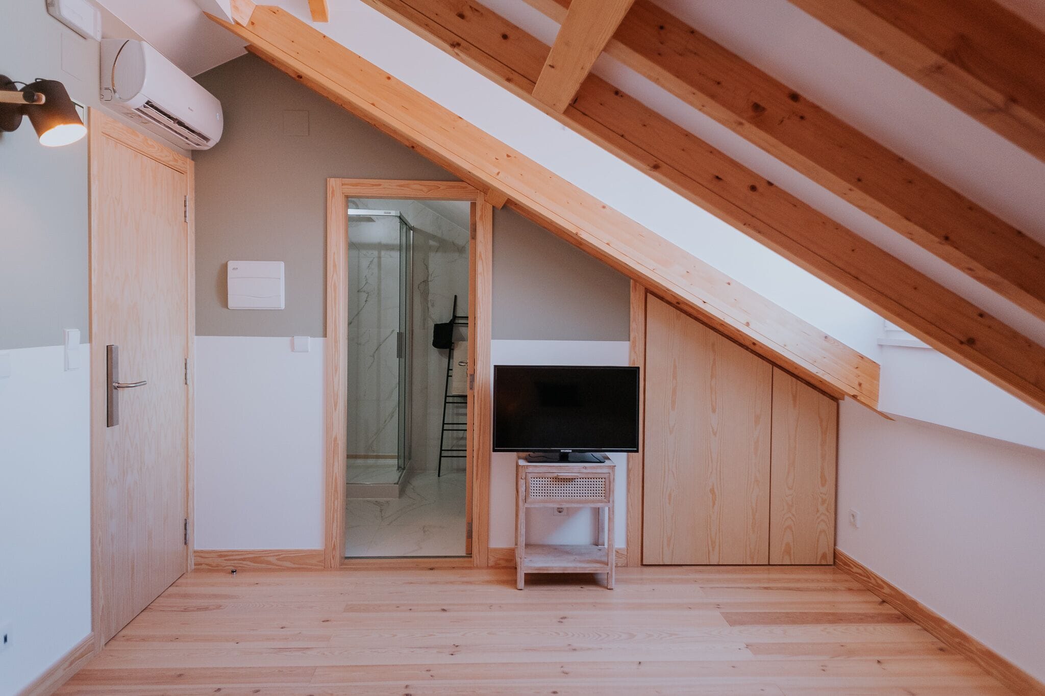 Una habitación pequeña en el ático, con techo inclinado, suelo de madera, un televisor sobre un mueble bajo y una puerta que da al baño.