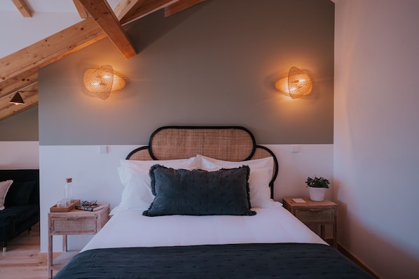 Un dormitorio con cama, dos mesitas de noche, una lámpara auxiliar, una planta en maceta y un techo inclinado con vigas a la vista.