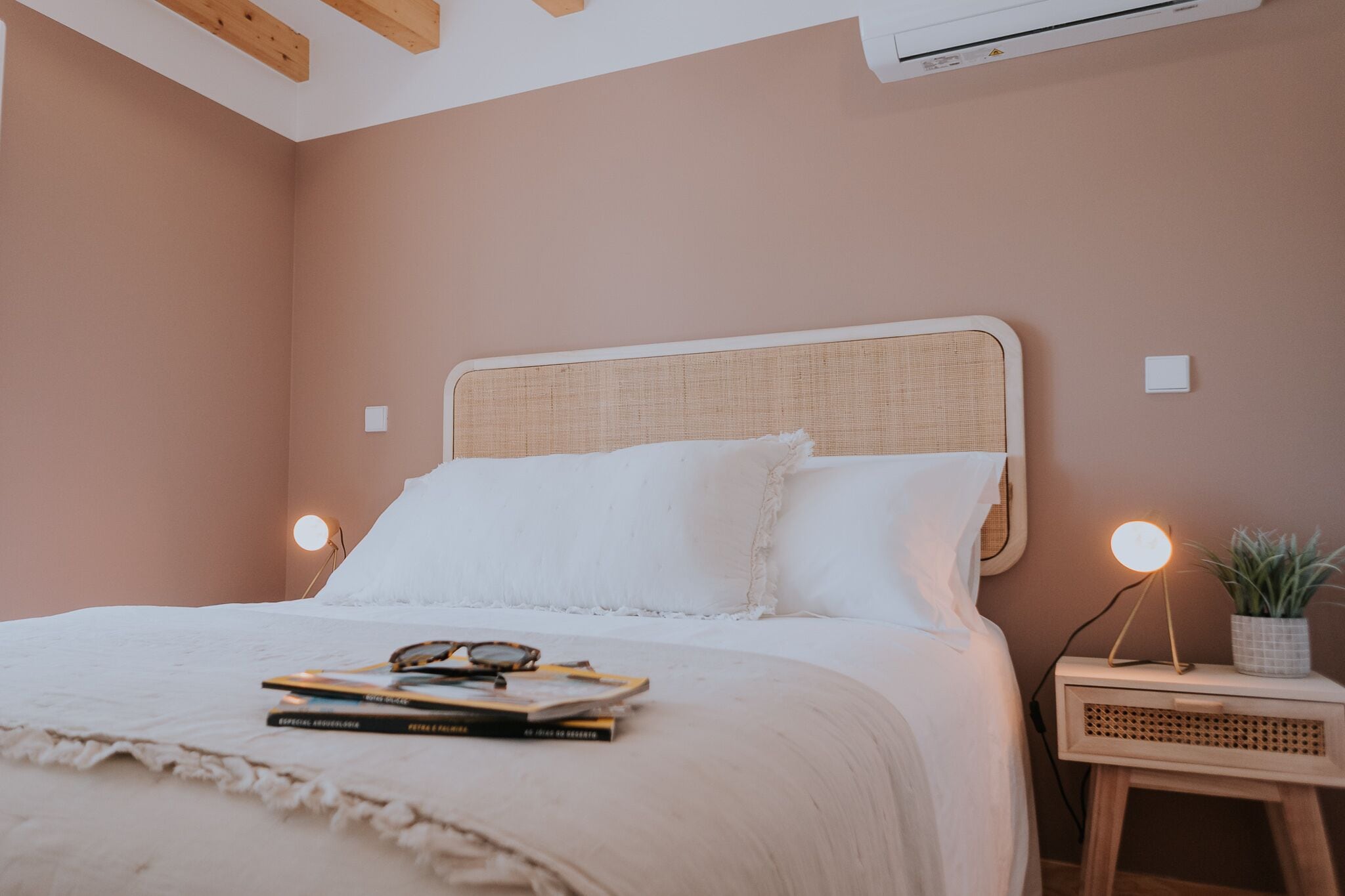 A bedroom with a bed, bedside lamps, a small table with a plant, and a wall-mounted air conditioner.