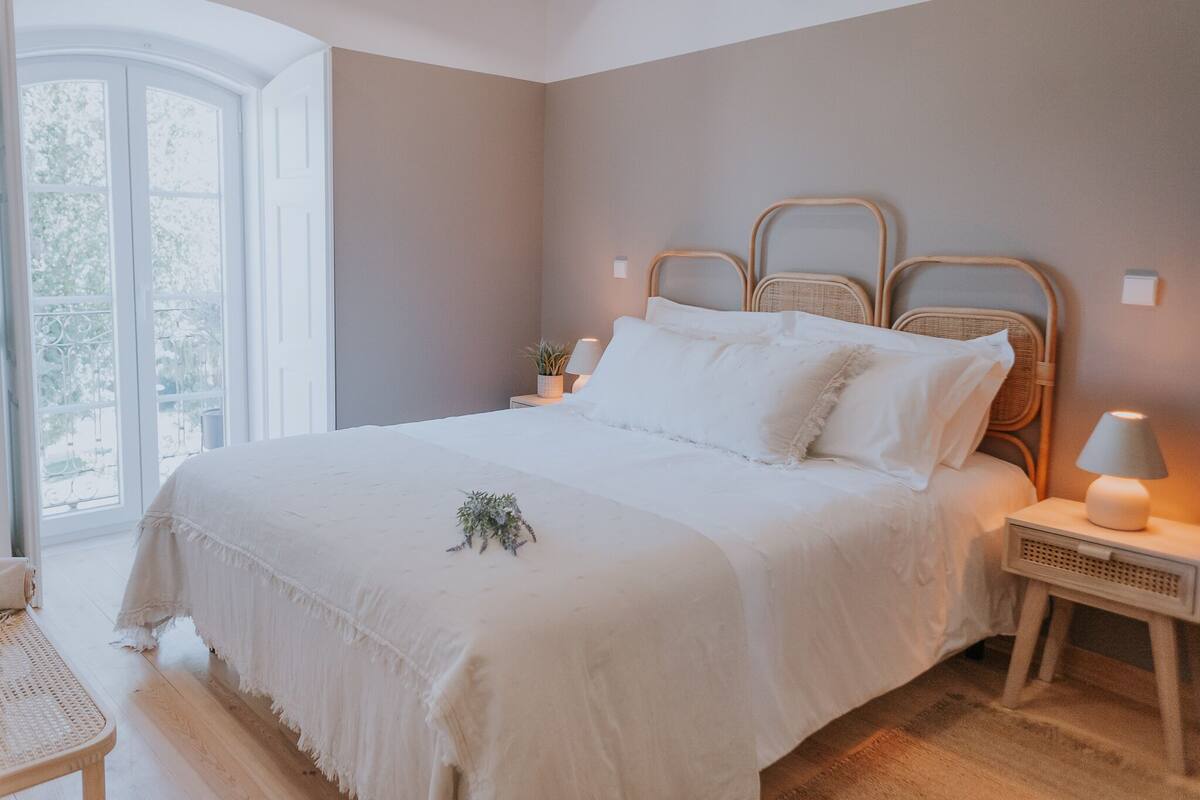 A bedroom with a large bed, a bedside table with a lamp, and a window with a view of greenery.