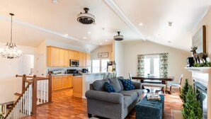 A spacious living area with a grey sofa and ottoman, wooden flooring, and a kitchen in the background. - The Wild Rose ~ A Custom Glam Cottage with Theater! In Historic Old St Charles (Saint Charles)