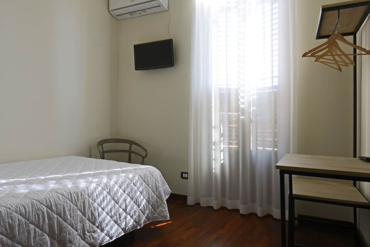 A bedroom with a bed, a chair, a table, a TV, and a window with curtains.